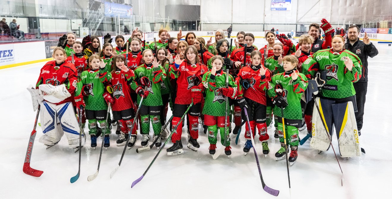 Group photo of the junior team in full kit on the ice rink, holding their hands up in the National Lottery crossed-finger pose.