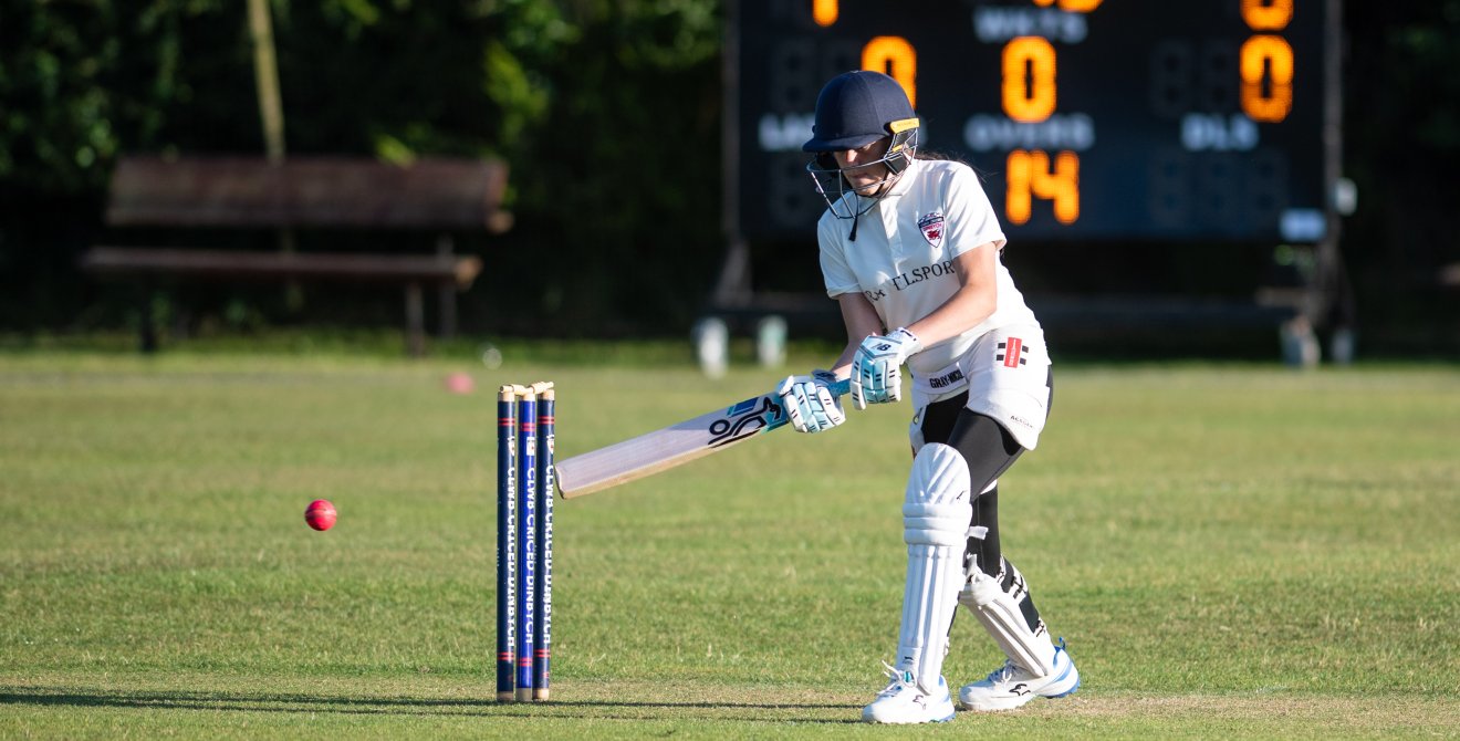 A girl playing cricket hits a shot with an electronic scoreboard in the background