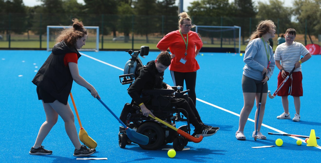People playing adapted hockey on a blue outdoor pitch, including a participant using a powered wheelchair