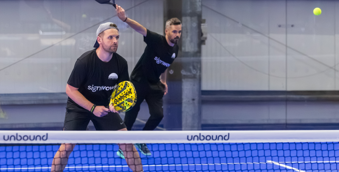 Two deaf padel players stand ready behind the net on an indoor blue court, holding rackets and watching the ball during a doubles match.