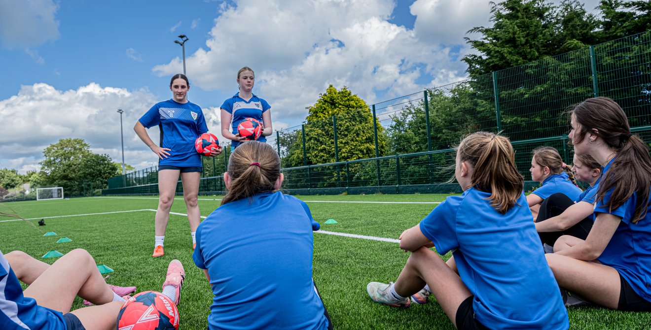 Group of young football players sitting on an outdoor artificial pitch, listening to two coaches holding footballs during a training session.