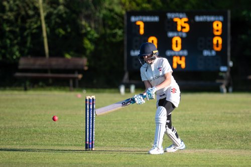 A girl playing cricket hits a shot with an electronic scoreboard in the background