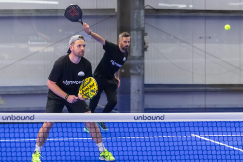 Two deaf padel players stand ready behind the net on an indoor blue court, holding rackets and watching the ball during a doubles match.