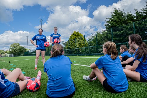 Group of young football players sitting on an outdoor artificial pitch, listening to two coaches holding footballs during a training session.