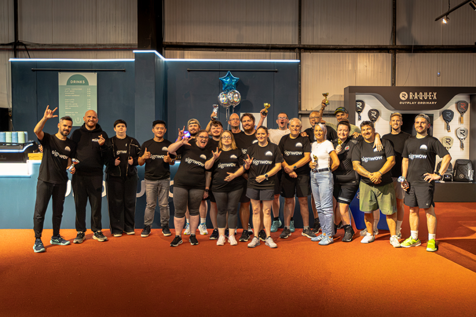 Deaf padel participants pose together indoors for a group photo. Some are lifting trophies